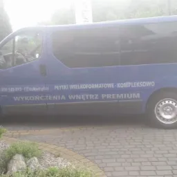 Niebieski bus z napisem 'Płytki wielkoformatowe - kompleksowo, wykończenia wnętrz premium' zaparkowany na brukowanym podjeździe, widoczne krzewy i budynek w tle, pochmurny dzień.