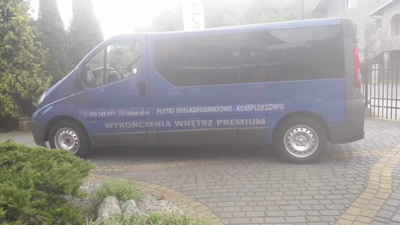 Niebieski bus z napisem 'Płytki wielkoformatowe - kompleksowo, wykończenia wnętrz premium' zaparkowany na brukowanym podjeździe, widoczne krzewy i budynek w tle, pochmurny dzień.