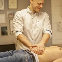 Mężczyzna w białej koszuli wykonuje masaż brzucha u pacjenta leżącego na kozetce w gabinecie, na ścianie wiszą dyplomy i zegar.