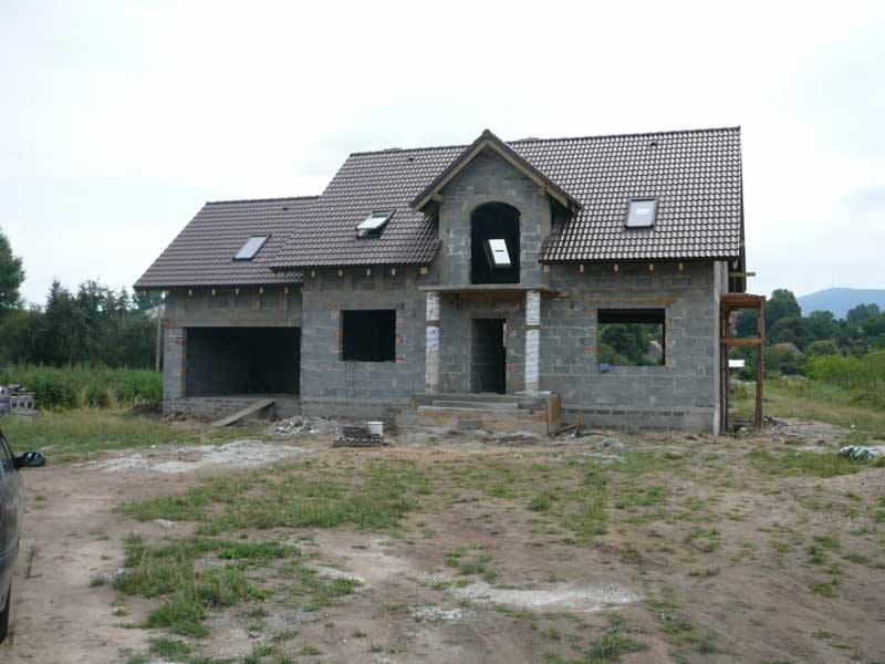 Budowa domu jednorodzinnego w stanie surowym otwartym, widoczna konstrukcja z bloczków betonowych, brązowa dachówka i otwarte okna dachowe, na pierwszym planie teren budowy z częściowo wykończonym...