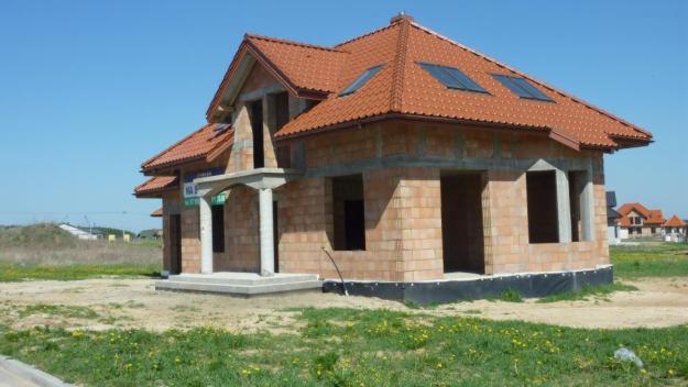 Niedokończony dom jednorodzinny z czerwonym dachem w stanie surowym otwartym, widoczne cegły, betonowe słupy wejściowe i czarna folia izolacyjna przy gruncie.
