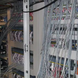 Rozbudowana szafa sterownicza z licznymi przewodami w kolorze szarym, białym, pomarańczowym i czerwonym, uporządkowane w wiązki, z widocznymi modułami sterującymi i zabezpieczeniami.