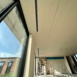 BARTOSZ SOPEL - Wnętrze w trakcie prac wykończeniowych: zamontowane płyty gipsowe na suficie z widocznymi szynami wentylacyjnymi, drabiny, duże okno z widokiem na budynek.