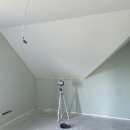 BARTOSZ SOPEL - Pokój na poddaszu po malowaniu ścian na pastelowy odcień zieleni, z białym sufitem i żółtą taśmą malarską wzdłuż krawędzi. Widoczny statyw oświetleniowy.