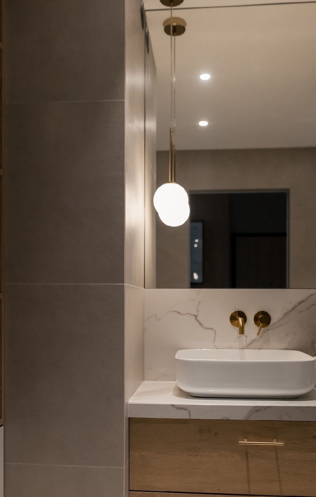 Nowoczesna łazienka z umywalką nablatową, marmurowym blatem i złotą armaturą. Minimalistyczna lampa wisząca z mleczną kulą. Szare płytki na ścianie.