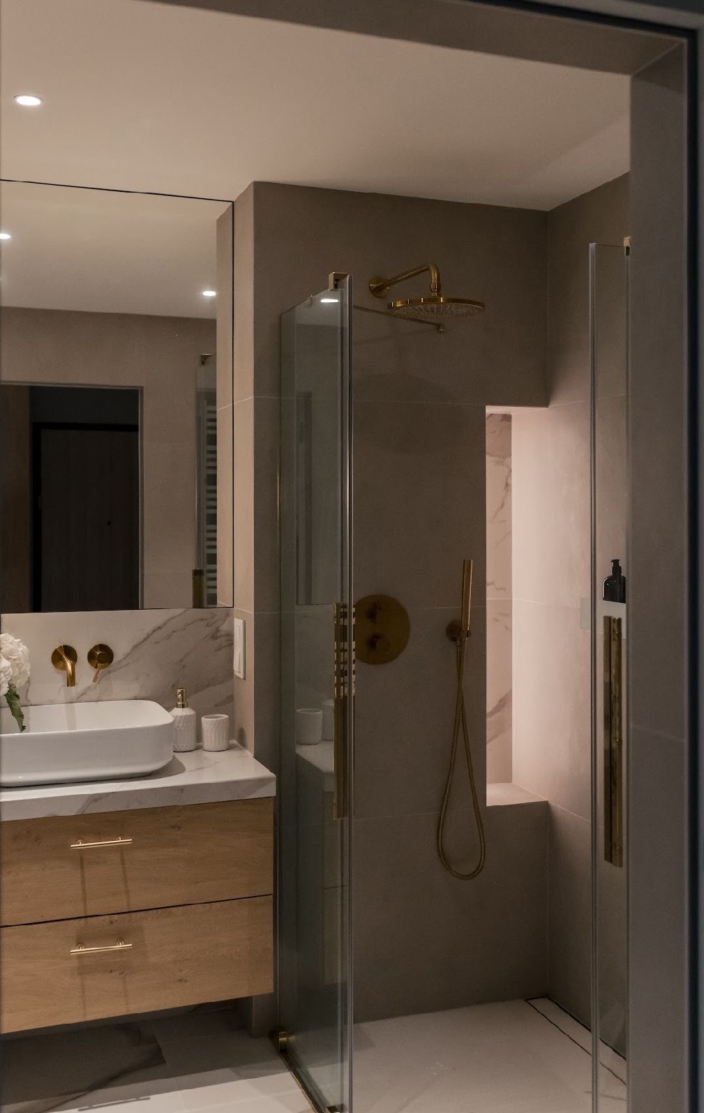 Nowoczesna łazienka z kabiną prysznicową typu walk-in, umywalką nablatową na drewnianej szafce i złotą armaturą. Minimalistyczny design, stonowana kolorystyka.