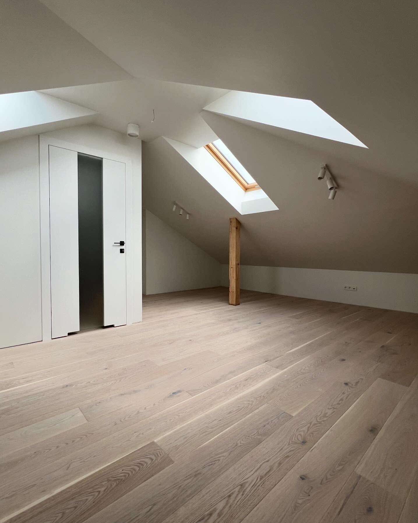 Jasne panele podłogowe w nowym pokoju na poddaszu z oknem dachowym i białymi drzwiami z czarnym paskiem. Widoczne gniazdka elektryczne i drewniany słup.