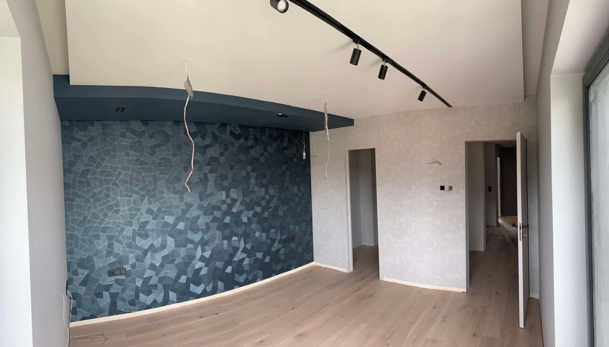 Wykończone wnętrze: parkiet, ściana z geometrycznym wzorem w odcieniach szarości i błękitu, sufit z oświetleniem szynowym. Widoczne otwarte drzwi i fragment okna.