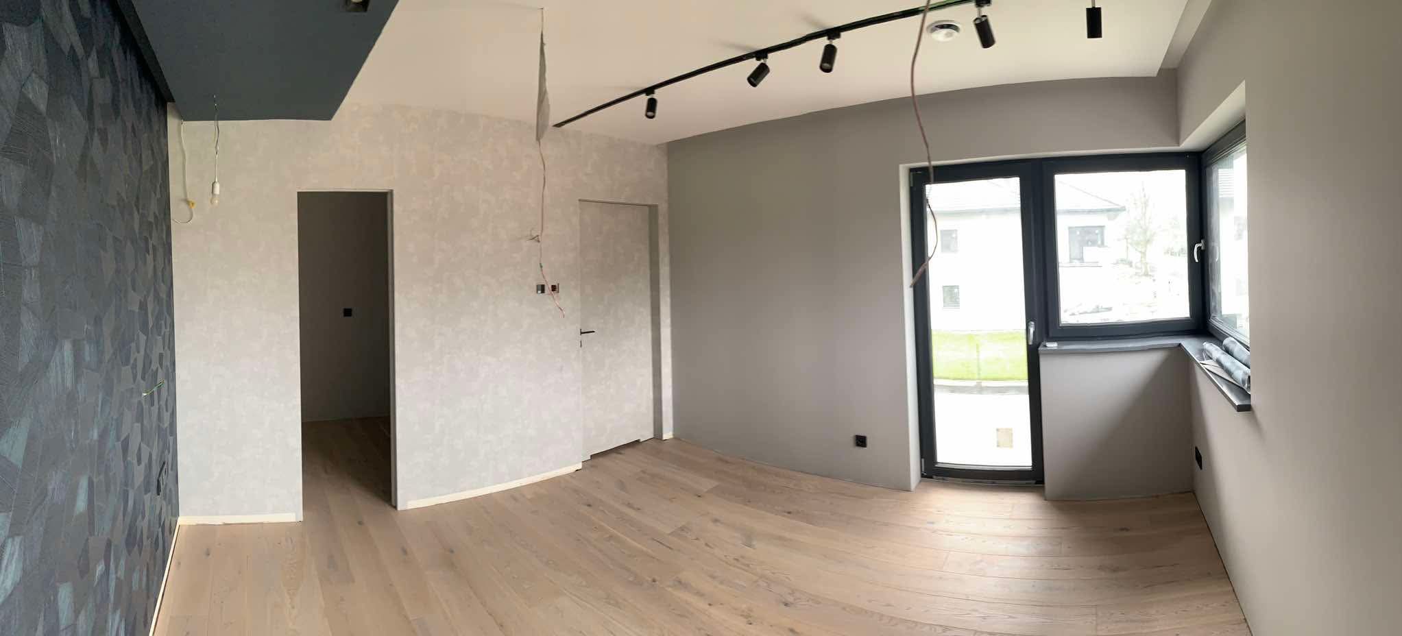 Jasne panele podłogowe w przestronnym pokoju z szarą ścianą, oknem i dekoracyjną, ciemną ścianą z geometrycznym wzorem. Widoczne niedokończone prace elektryczne.