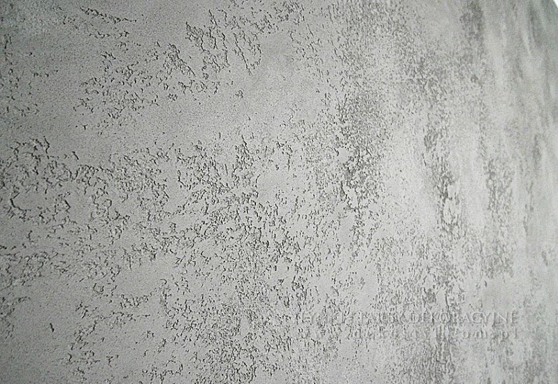 Szara ściana z wyraźną, nieregularną fakturą dekoracyjną, imitującą naturalny kamień lub beton. Widoczne drobne nierówności i cieniowania podkreślające strukturę.