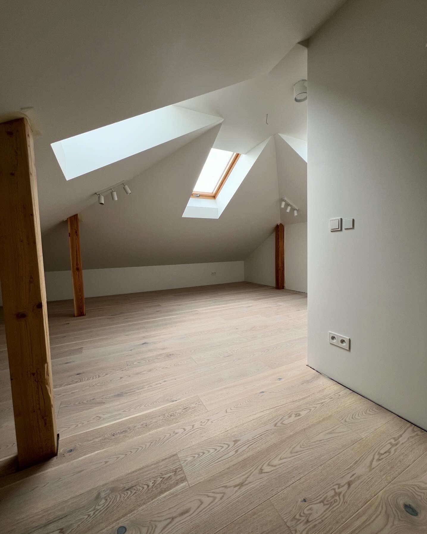 Jasne panele podłogowe w nowym, przestronnym pokoju na poddaszu z drewnianymi belkami i oknami dachowymi. Świeżo pomalowane ściany i widoczne gniazdka elektryczne.