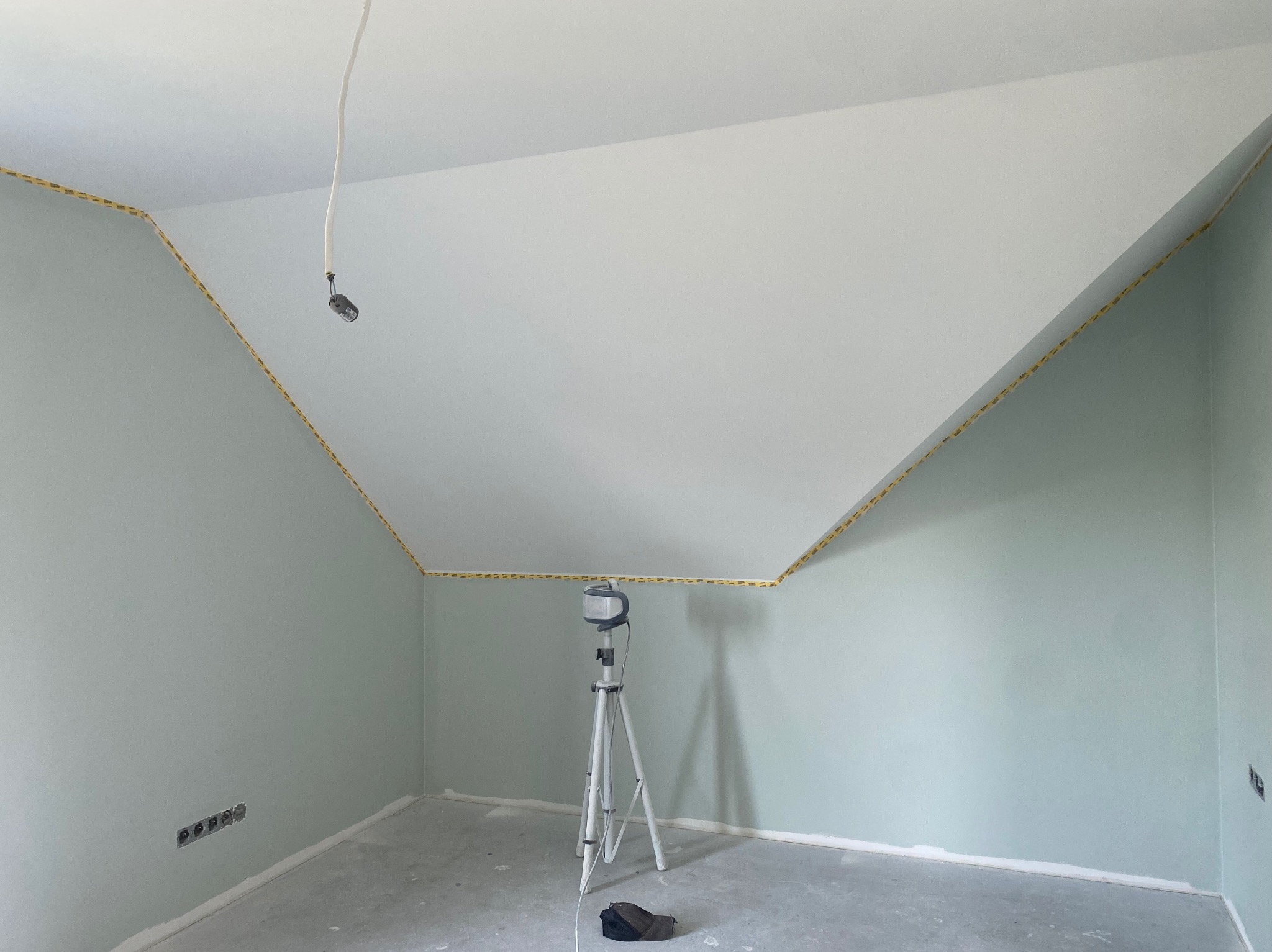 Pokój na poddaszu po malowaniu ścian na pastelowy odcień zieleni, z białym sufitem i żółtą taśmą malarską wzdłuż krawędzi. Widoczny statyw oświetleniowy.