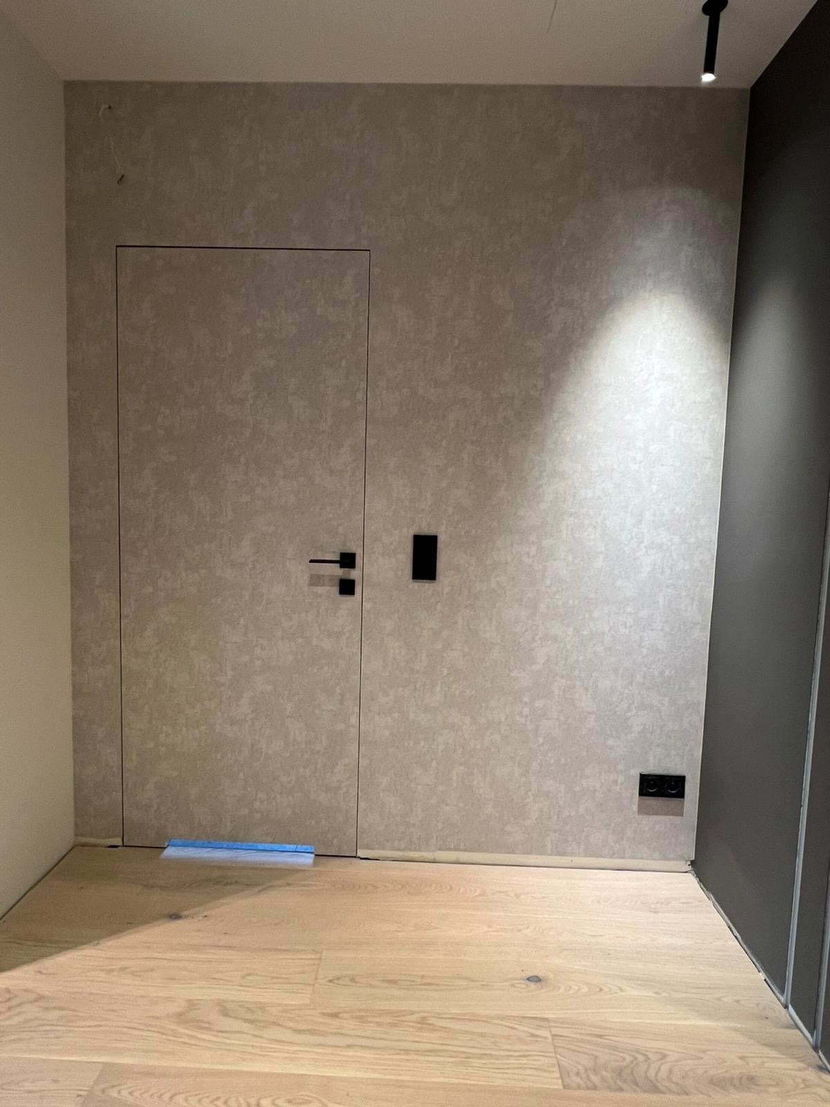 Ściana z ukrytymi drzwiami i gładką fakturą, czarne gniazdka i włączniki, drewniana podłoga, minimalistyczne oświetlenie punktowe. Nowoczesne wykończenie wnętrza.