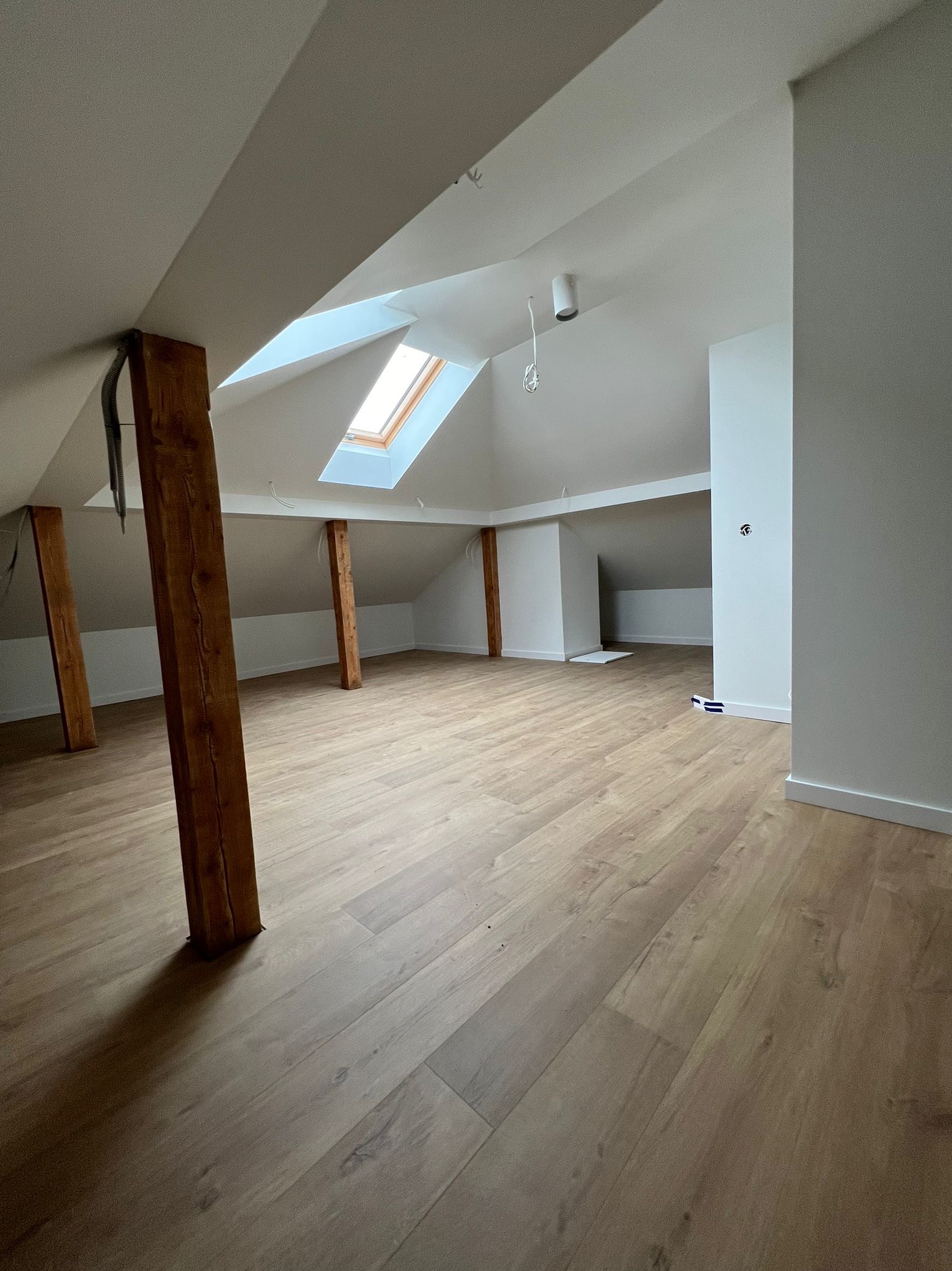 Jasne poddasze po remoncie z drewnianymi belkami, oknem dachowym i panelami podłogowymi. Świeżo pomalowane ściany i sufit. Widoczne detale wykończeniowe.