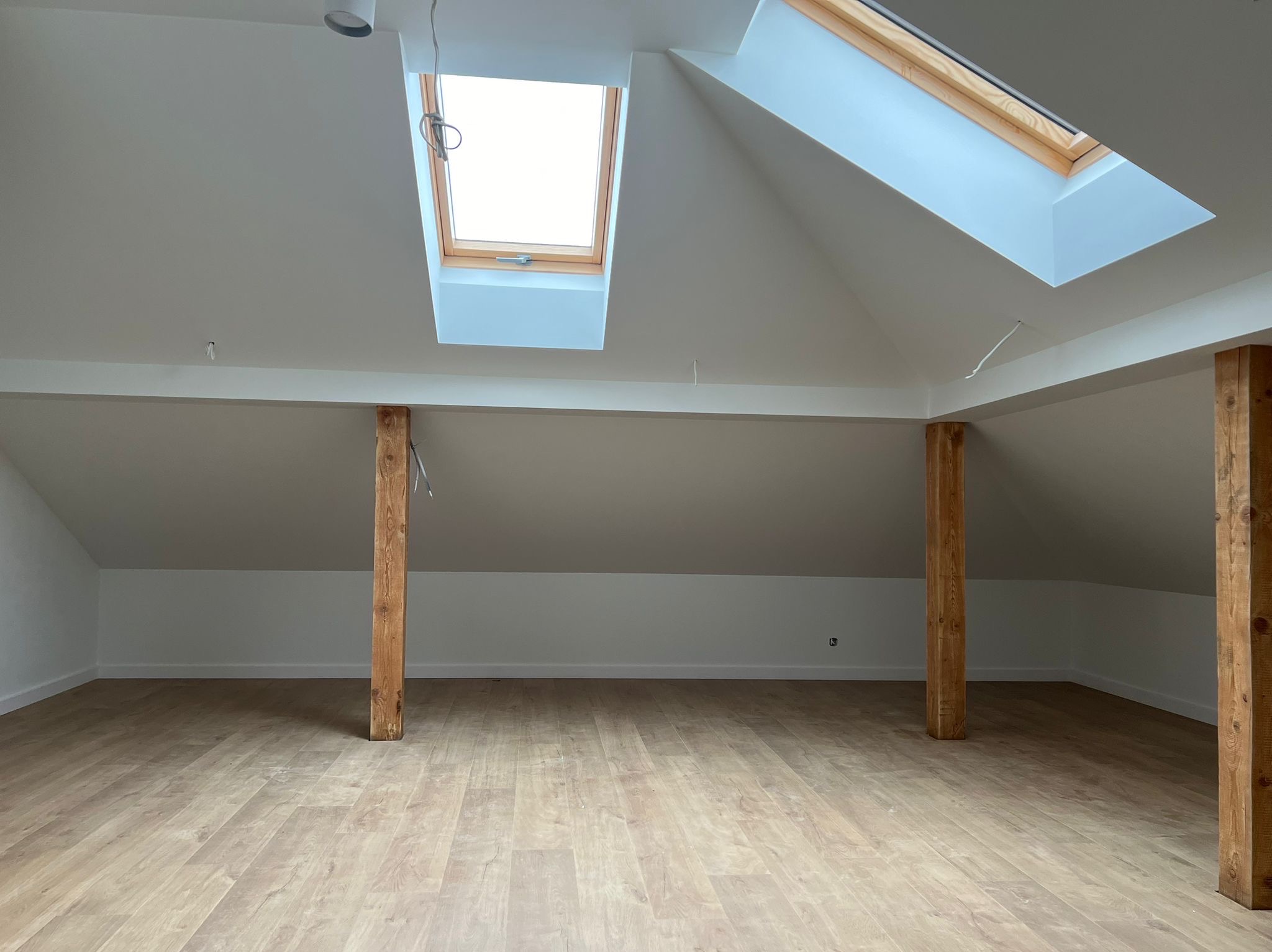 Jasne poddasze po remoncie: drewniane słupy, panele podłogowe i dwa okna dachowe w białym suficie. Ściany pomalowane na biało, widoczne kable.