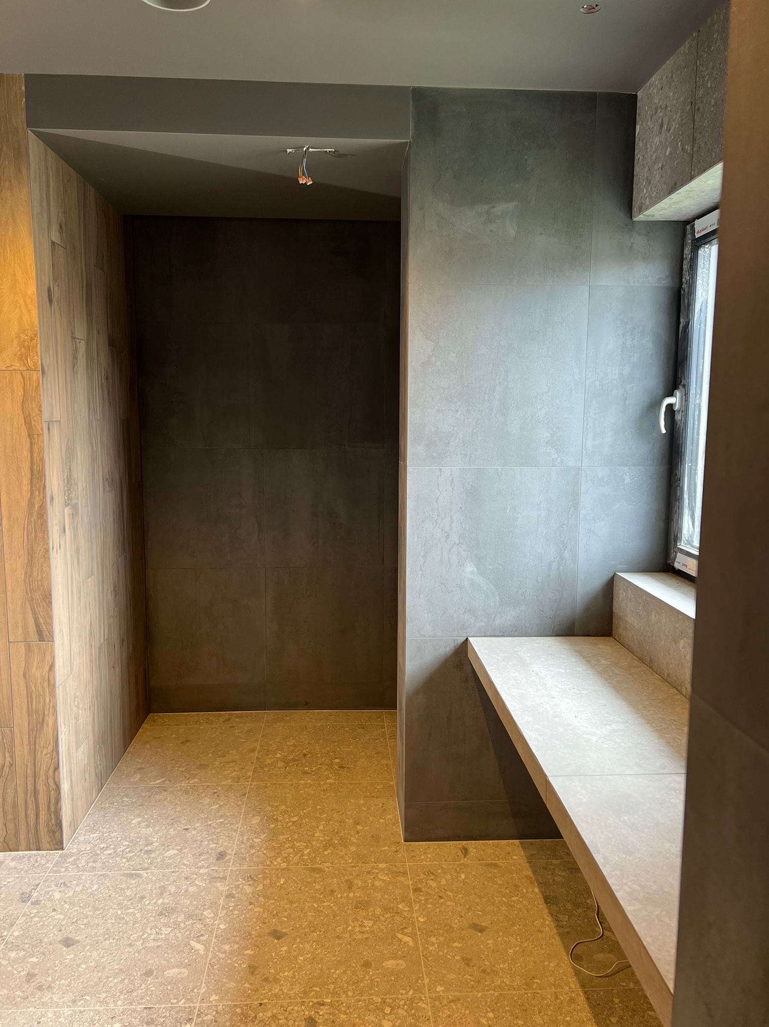 Wykończone wnętrze z szarymi płytkami na ścianach, terakotą na podłodze i drewnopodobną okładziną. Widoczny fragment okna i półka. Minimalistyczny design.