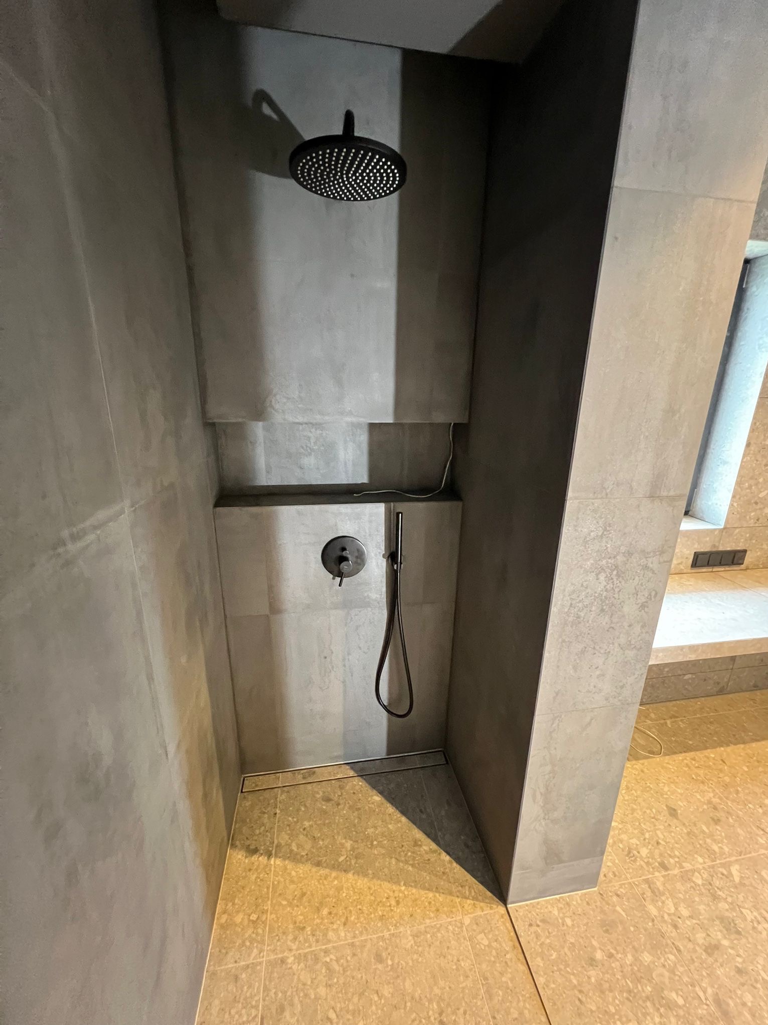 Nowoczesna kabina prysznicowa z szarą armaturą i płytkami w odcieniach szarości, z odpływem liniowym i półką na kosmetyki. Minimalistyczny design.