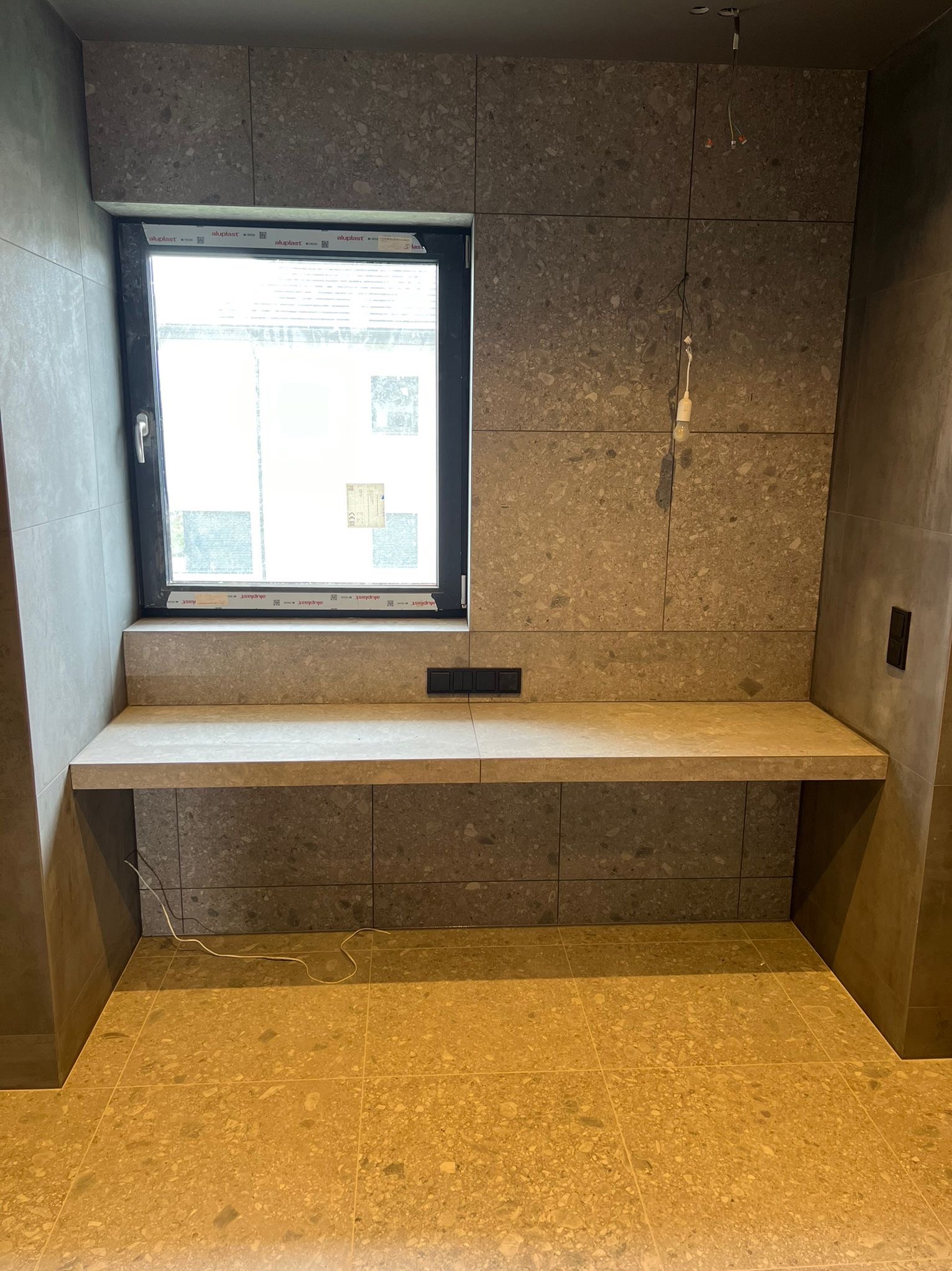 Wykończona wnęka okienna z blatem i płytkami w odcieniach szarości i beżu. Widoczne gniazdka elektryczne i fragment instalacji oświetleniowej.