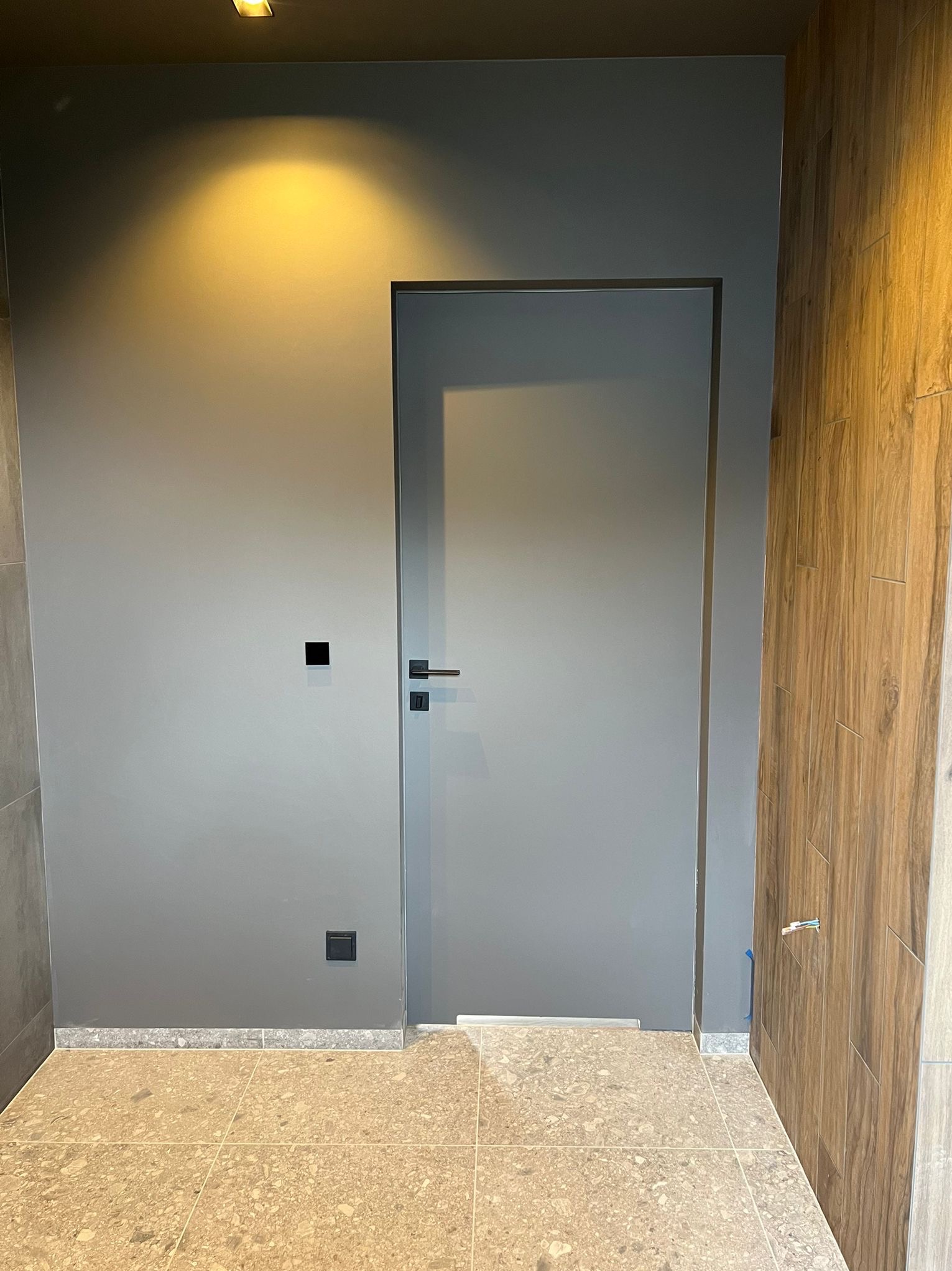 Szara ściana z drzwiami w minimalistycznym stylu, widoczny fragment drewnianej okładziny ściany i terakota na podłodze. Nowoczesne wykończenie wnętrza.