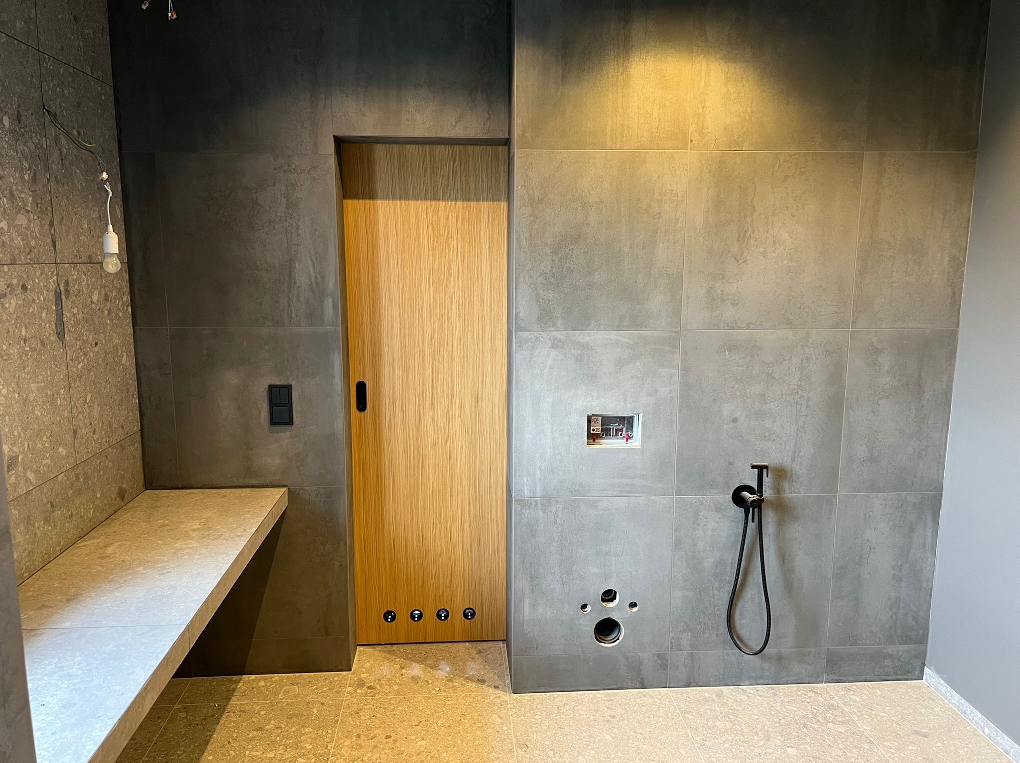 Szara łazienka z drewnianymi drzwiami i elementami armatury w trakcie montażu. Widoczne otwory na rury i instalacje, surowy stan wykończenia.