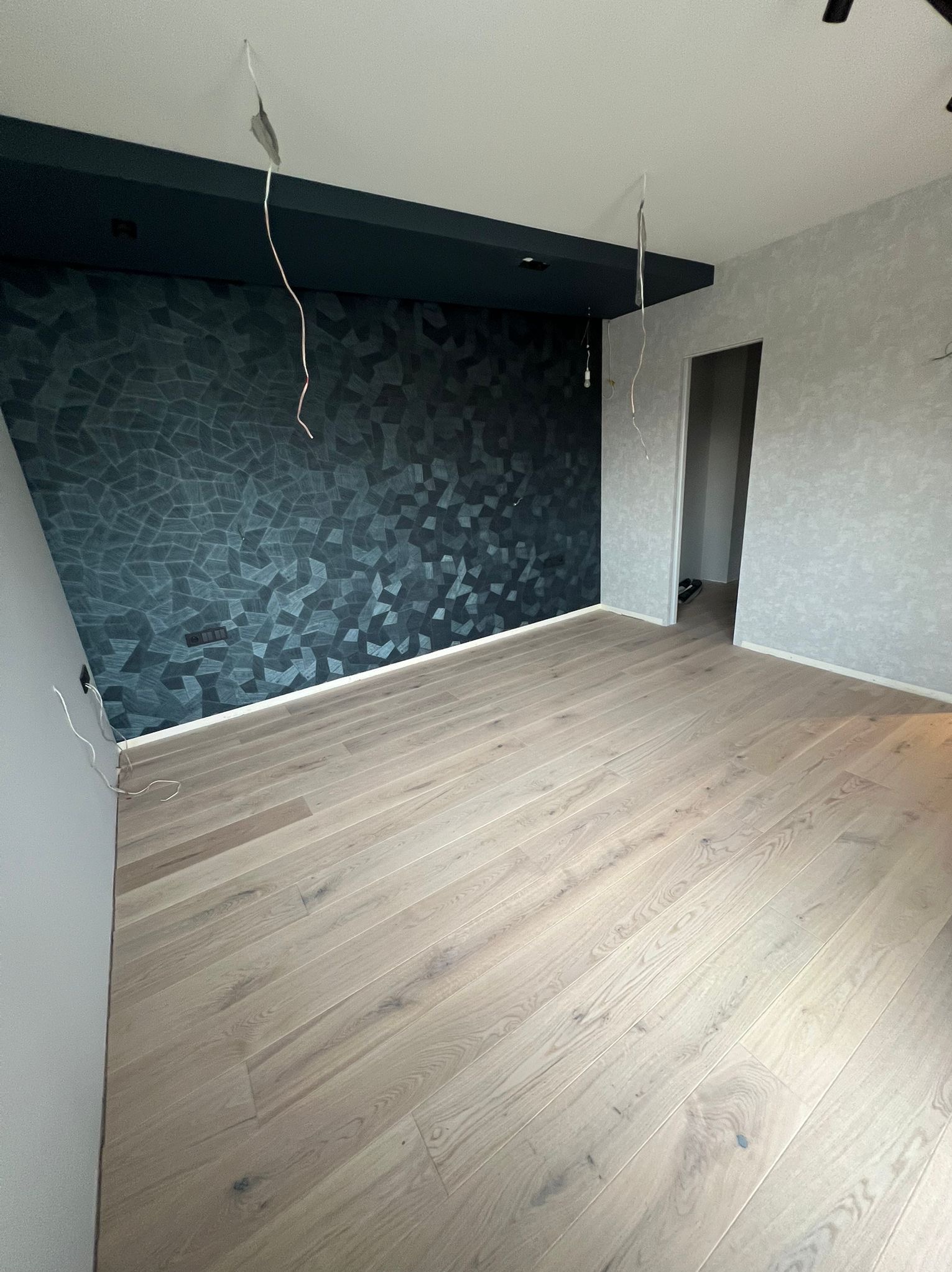 Jasne panele podłogowe w nowocześnie wykończonym pokoju z ciemną ścianą w geometryczne wzory i szarą ścianą z efektem dekoracyjnym. Widoczne kable wystające z sufitu.