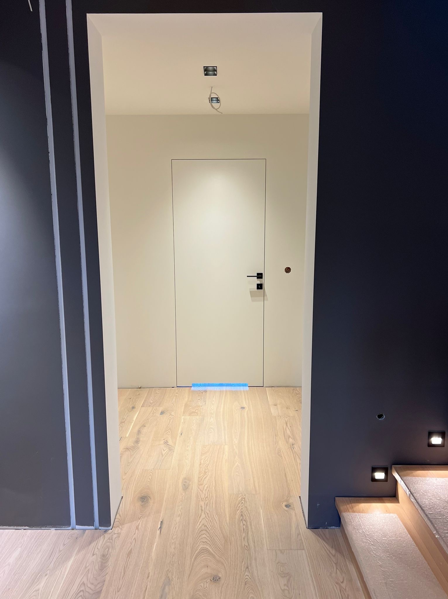 Nowoczesne wnętrze: białe drzwi z czarną klamką, drewniana podłoga, ciemna ściana z oświetleniem punktowym. Widok z korytarza.