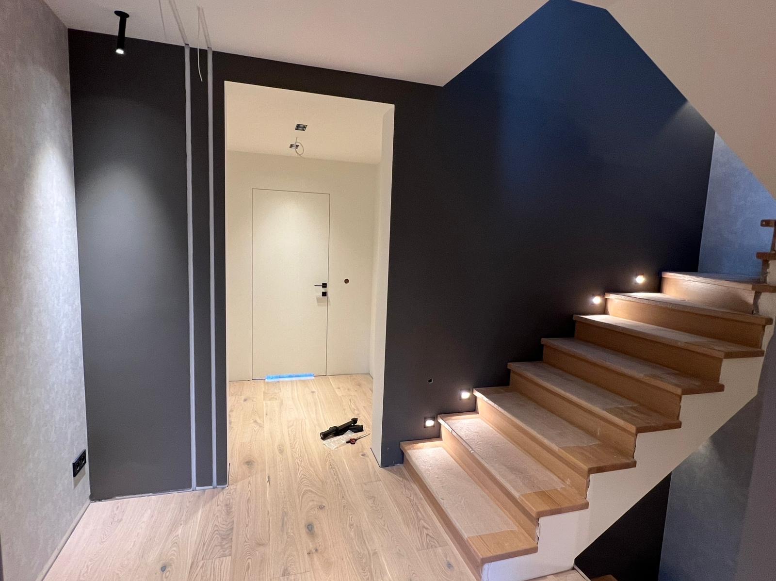 Nowoczesne wnętrze: drewniane schody z podświetleniem, ciemnoszara ściana i jasne panele podłogowe. Widoczne narzędzia sugerują prace wykończeniowe w toku.