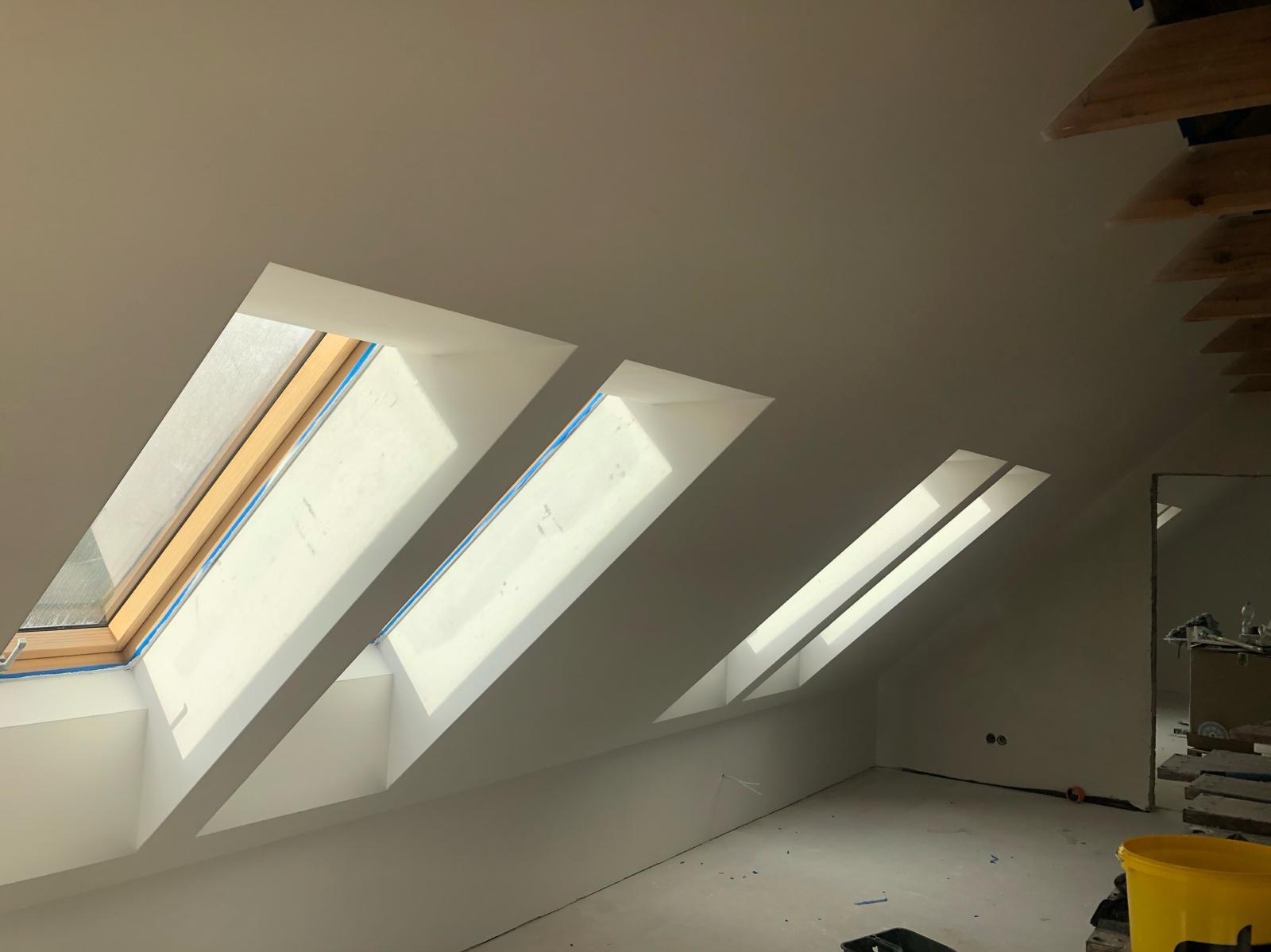 Poddasze po gładziowaniu i malowaniu na biało, z trzema oknami dachowymi, zabezpieczonymi folią malarską. Widoczne ślady prac wykończeniowych i fragmenty konstrukcji dachu.