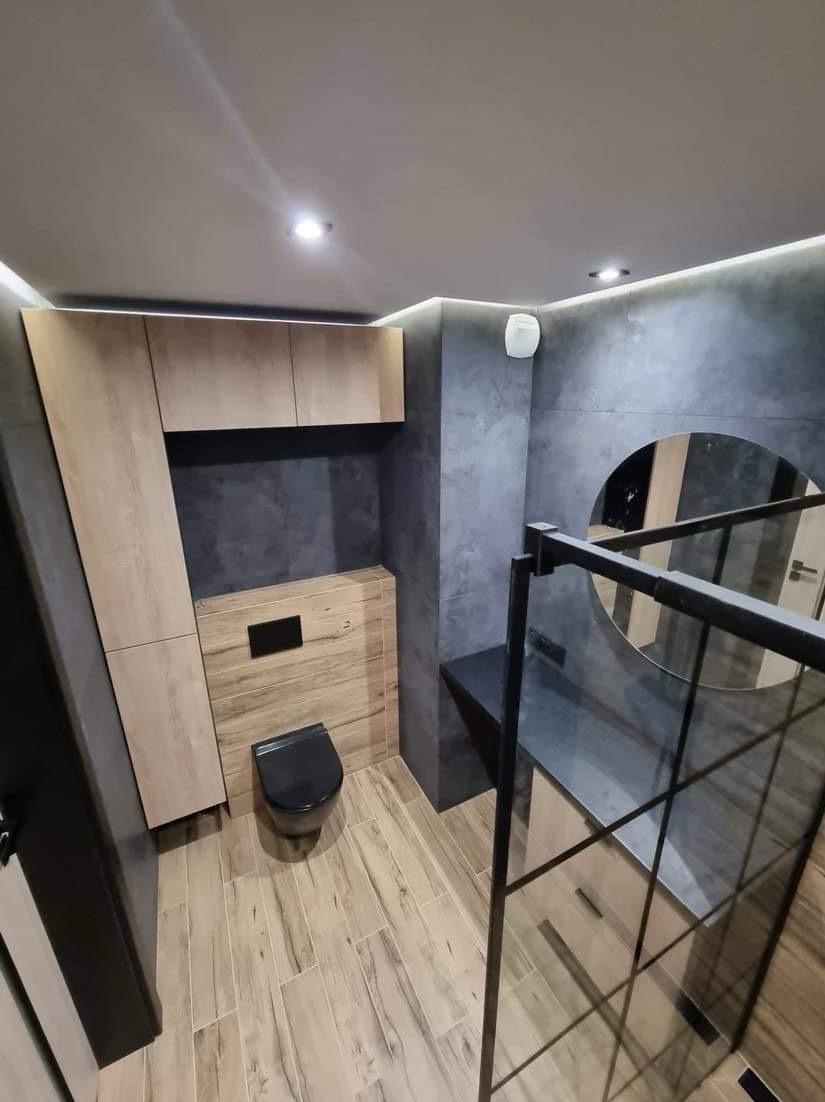 Nowoczesna łazienka po remoncie: drewnopodobne płytki, szare ściany z efektem betonu, czarna armatura i kabina prysznicowa z okrągłym lustrem. Oświetlenie LED.