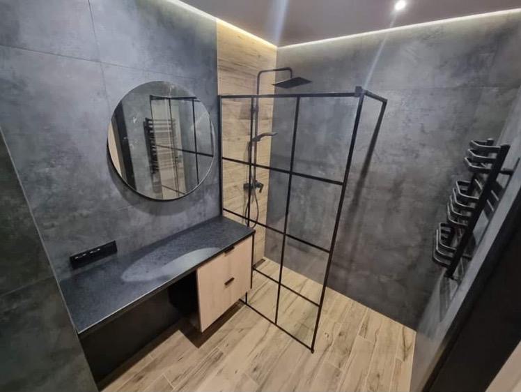 Nowoczesna łazienka z szarą płytką imitującą beton, drewnopodobną podłogą i kabiną prysznicową typu walk-in z czarnymi profilami. Okrągłe lustro nad umywalką.