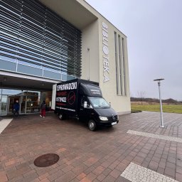 WHM Transport - Czarny bus z reklamą firmy przeprowadzkowej zaparkowany przed nowoczesnym budynkiem biblioteki. Widoczni ludzie wchodzący do budynku. Pochmurne niebo.