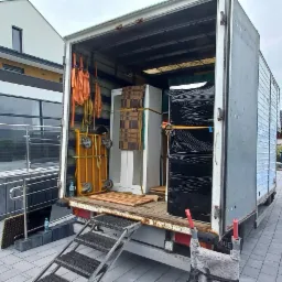 Otwarta paka samochodu dostawczego z rampą załadowczą, zabezpieczone meble gotowe do transportu, widok na nowoczesny budynek z rampą dla wózków inwalidzkich i parkingiem dla niepełnosprawnych.