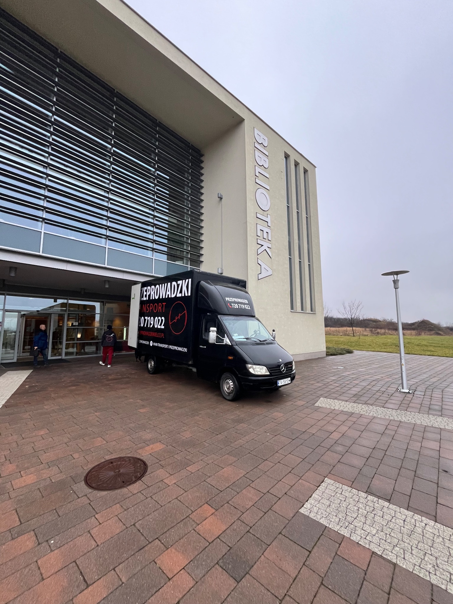 Czarny bus z reklamą firmy przeprowadzkowej zaparkowany przed nowoczesnym budynkiem biblioteki. Widoczni ludzie wchodzący do budynku. Pochmurne niebo.