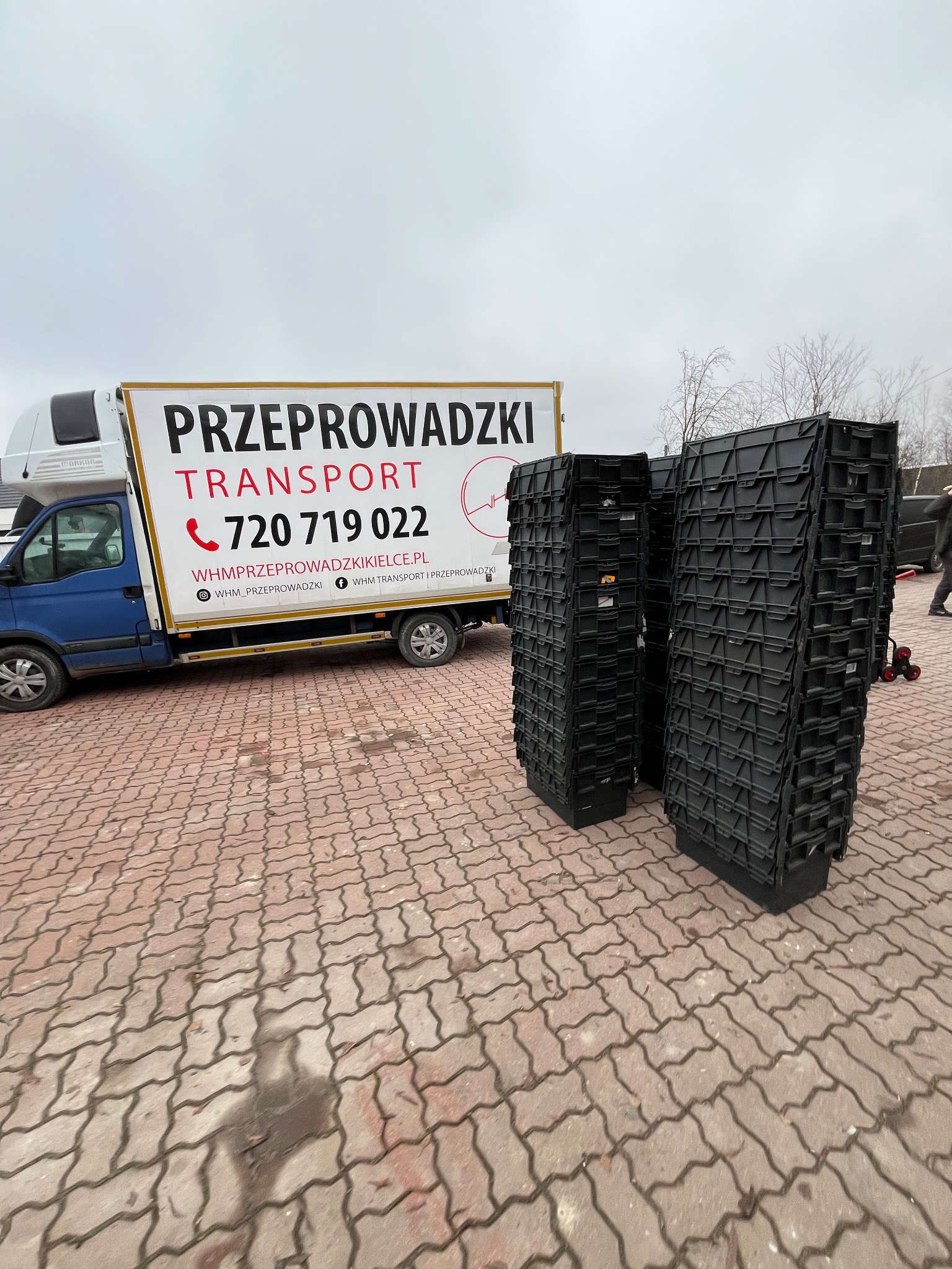 Ciężarówka z napisem 'Przeprowadzki Transport' i sterty czarnych pojemników na tle kostki brukowej. Widoczny numer telefonu i adres strony internetowej.