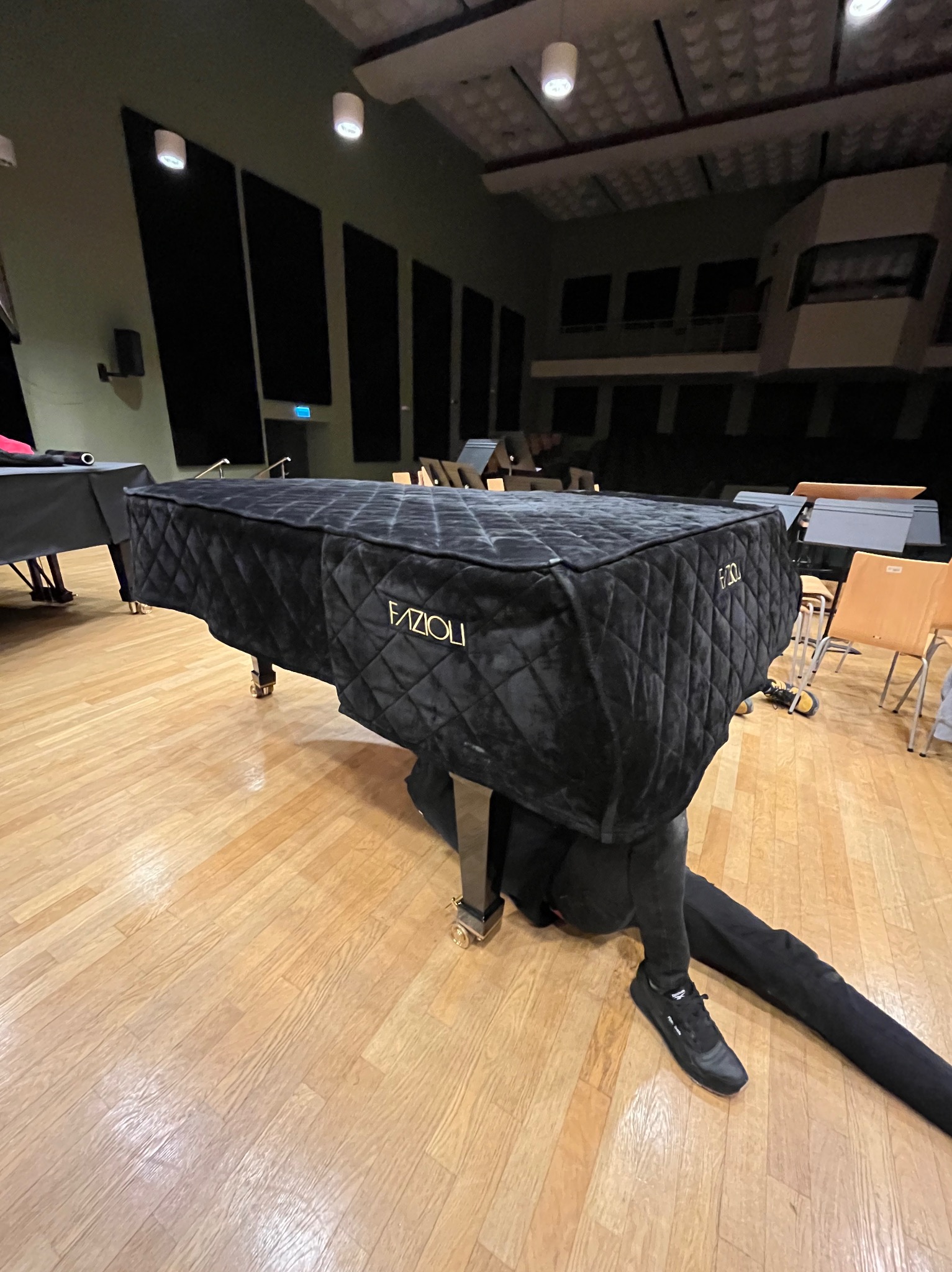 Czarny, pikowany pokrowiec na fortepian marki Fazioli w sali koncertowej. Osoba w czarnym ubraniu leży pod instrumentem, prawdopodobnie podczas transportu lub ustawiania.