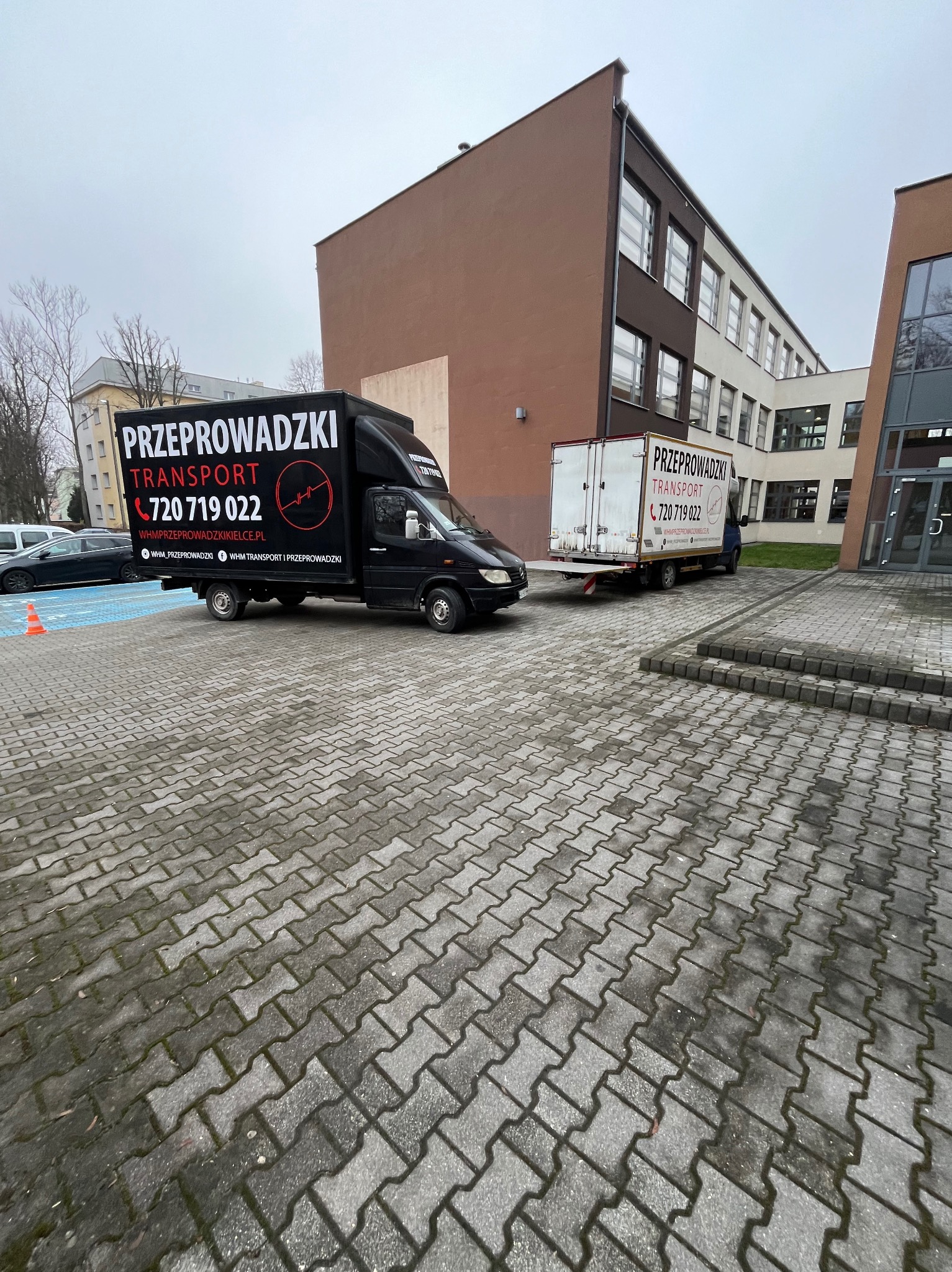 Dwa busy firmy przeprowadzkowej zaparkowane na brukowanym placu przed budynkiem. Na busach widoczne logo i numer telefonu. Jeden z busów ma otwartą platformę załadunkową.