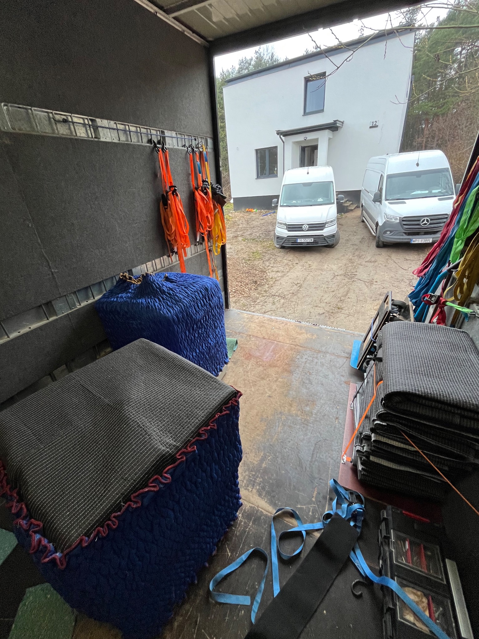 Wnętrze busa transportowego z zabezpieczonymi meblami (pufy, materace) i pasami mocującymi. Na zewnątrz widoczne dwa busy i budynek mieszkalny.