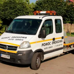 Biała laweta Opel Movano z napisem 'Pomoc Drogowa Auto-Serwis' i pomarańczowymi światłami ostrzegawczymi na dachu, zaparkowana na kostce brukowej z zielonymi krzewami w tle.