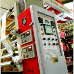 Maszyna drukarska z panelem sterowania, widoczne rolki z folią i wyświetlacze cyfrowe, w tle fragment hali produkcyjnej.