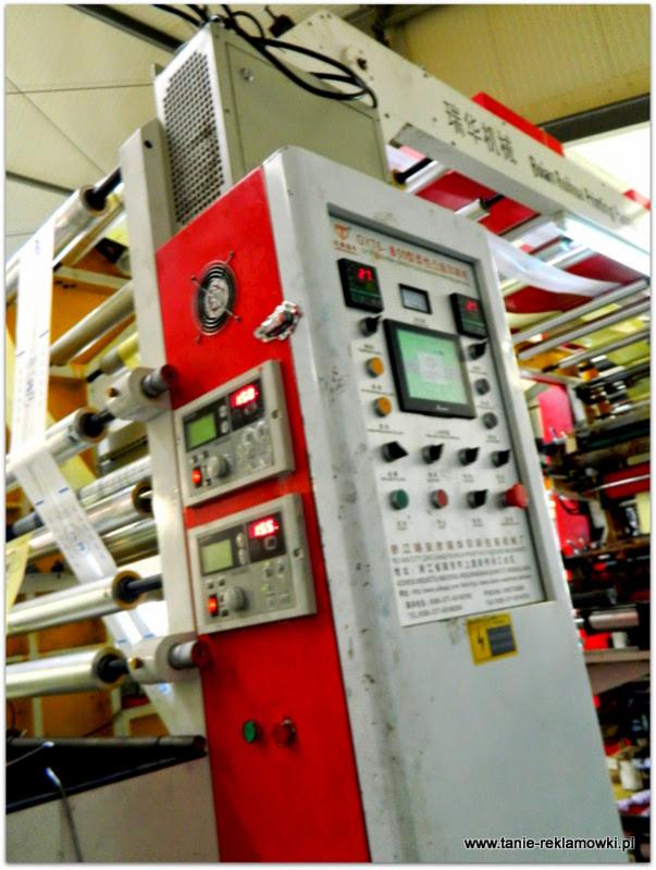 Maszyna drukarska z panelem sterowania, widoczne rolki z folią i wyświetlacze cyfrowe, w tle fragment hali produkcyjnej.