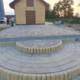 Ukończona nawierzchnia z kostki brukowej w dwóch odcieniach, ułożona w okrągłe schody prowadzące do podjazdu, z domkami w tle i rozproszonym światłem zachodzącego słońca.