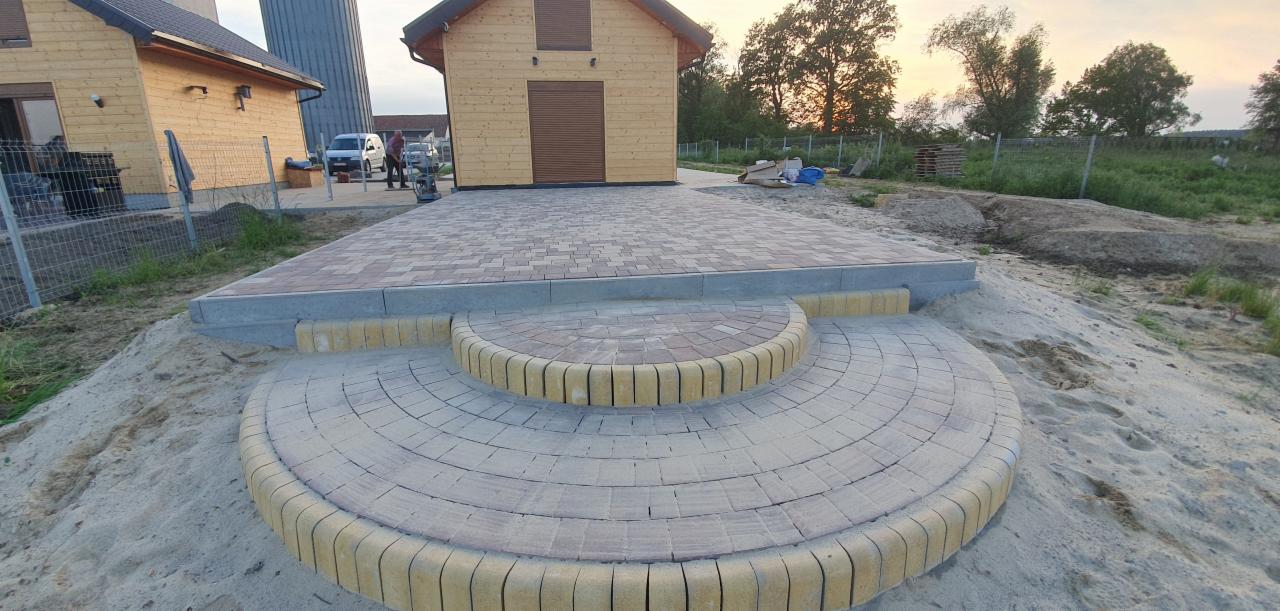 Ukończona nawierzchnia z kostki brukowej w dwóch odcieniach, ułożona w okrągłe schody prowadzące do podjazdu, z domkami w tle i rozproszonym światłem zachodzącego słońca.