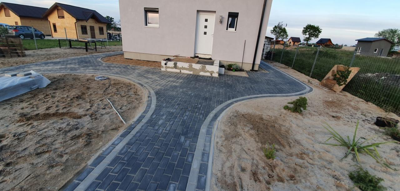 Nowo ułożona, ciemnoszara kostka brukowa z jasnoszarą obwódką tworząca ścieżkę do domu; widoczne pozostałości piasku i przygotowane miejsce na trawnik.