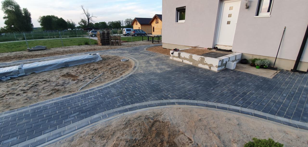 Nowo ułożona, szara kostka brukowa tworząca ścieżkę i podjazd przed domem z białymi drzwiami wejściowymi, widoczne elementy przygotowania terenu, w tle drewniany dom i zieleń.