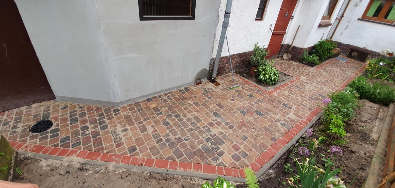 Ułożona z kostki brukowej ścieżka wokół domu, z obrzeżem z czerwonej kostki, prowadząca do drzwi i ogrodu z roślinami.