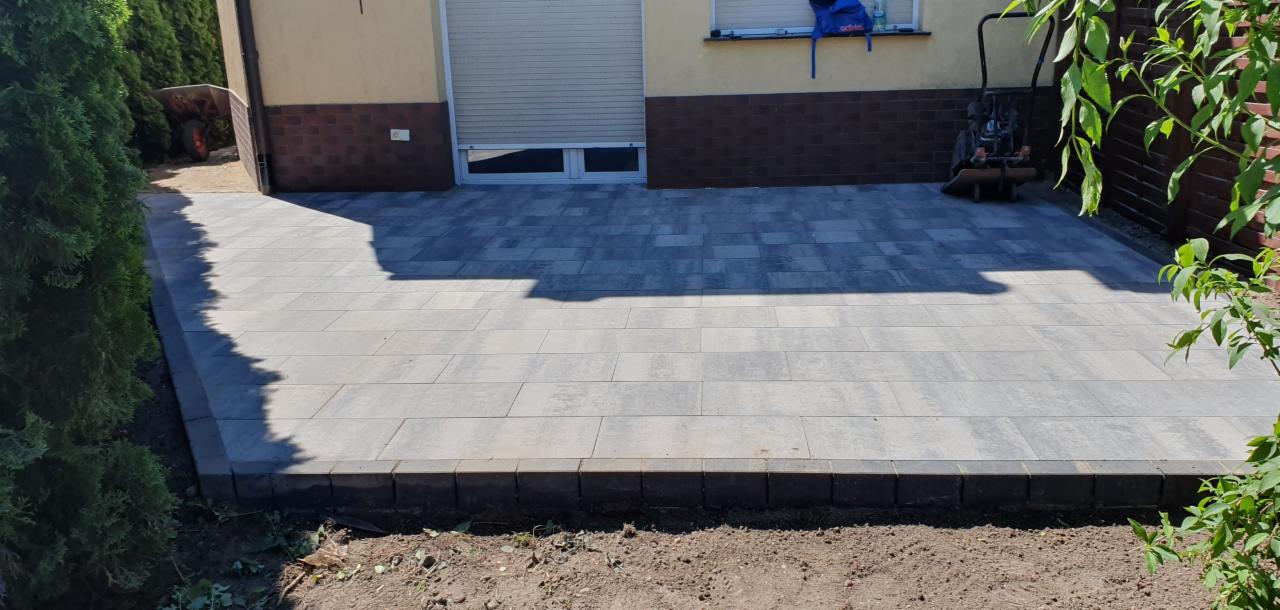 Nowo ułożony, szary chodnik z kostki brukowej, obramowany ciemniejszymi kostkami, przylegający do budynku z brązową elewacją i zielonymi krzewami po bokach. Na chodniku cień rzucany przez budynek.