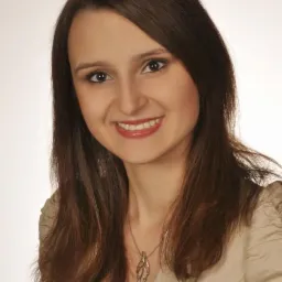 Uśmiechnięta brunetka w beżowej marynarce i z wisiorkiem w kształcie liścia na białym tle. Portret kobiety, nauczycielki lub tłumaczki.