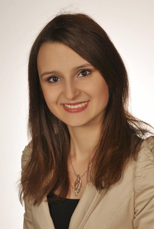 Uśmiechnięta brunetka w beżowej marynarce i z wisiorkiem w kształcie liścia na białym tle. Portret kobiety, nauczycielki lub tłumaczki.