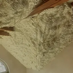 Poddasze z drewnianymi belkami pokryte grubą warstwą jasnej pianki izolacyjnej, widoczne metalowe druty i elementy mocujące, fragment okna i schodów w tle.