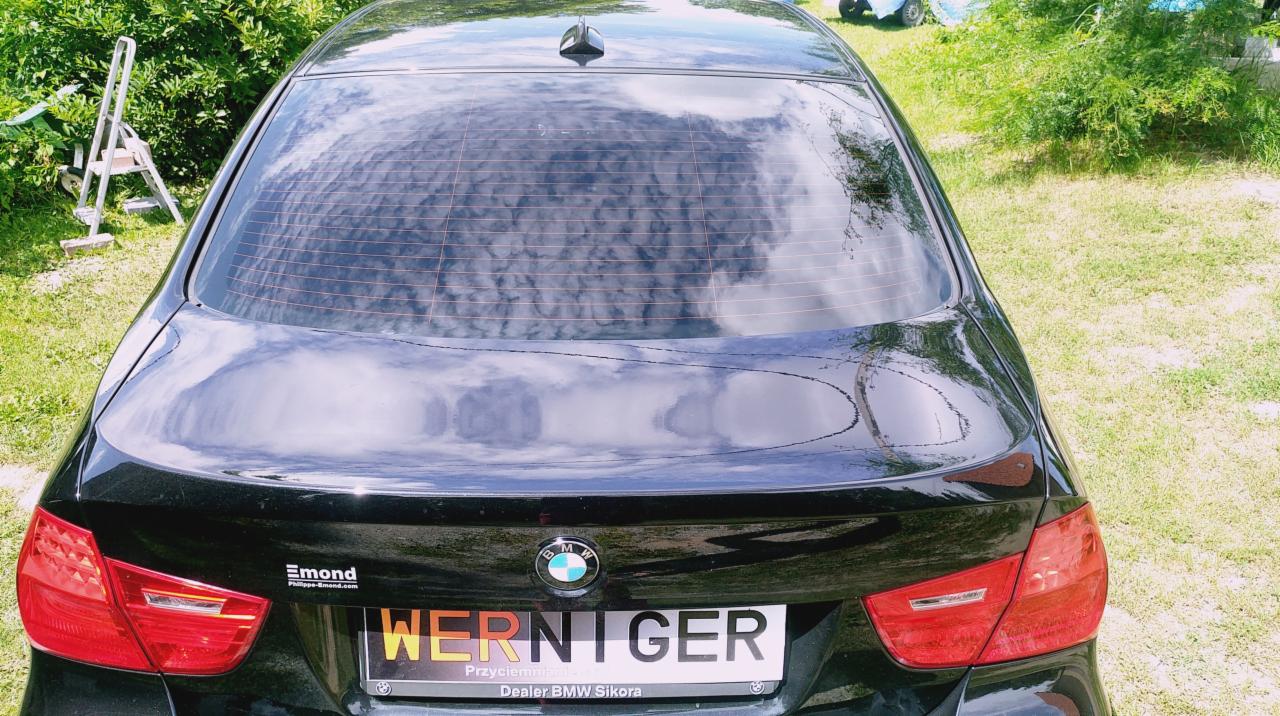 Czarne BMW z widocznym przyciemnionym tylnym oknem, odbijającym chmury, z tablicą rejestracyjną WERNIGER. W tle zieleń i drabina.